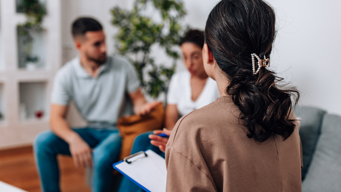 The camera focuses on the back of a female therapist's head. She is speaking with a couple who are sitting across from her.