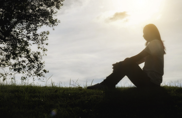 A grieving woman sits against the backdrop of a bright sky, her silhouette black in the middle of the day.