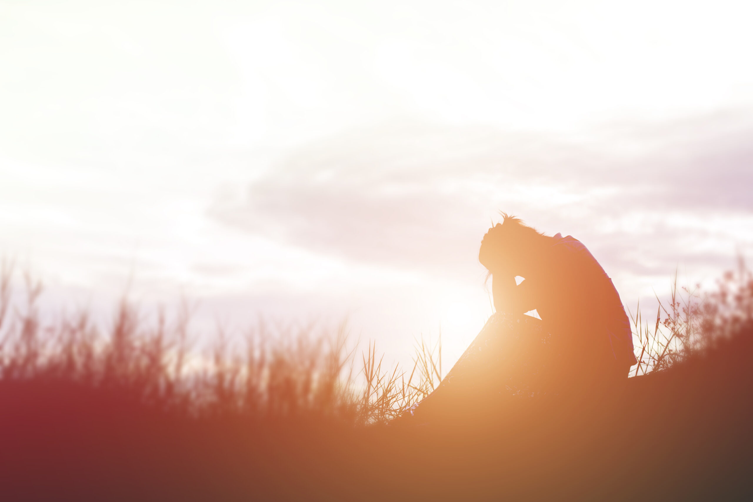 A grieving person sits alone in a field at sunset.