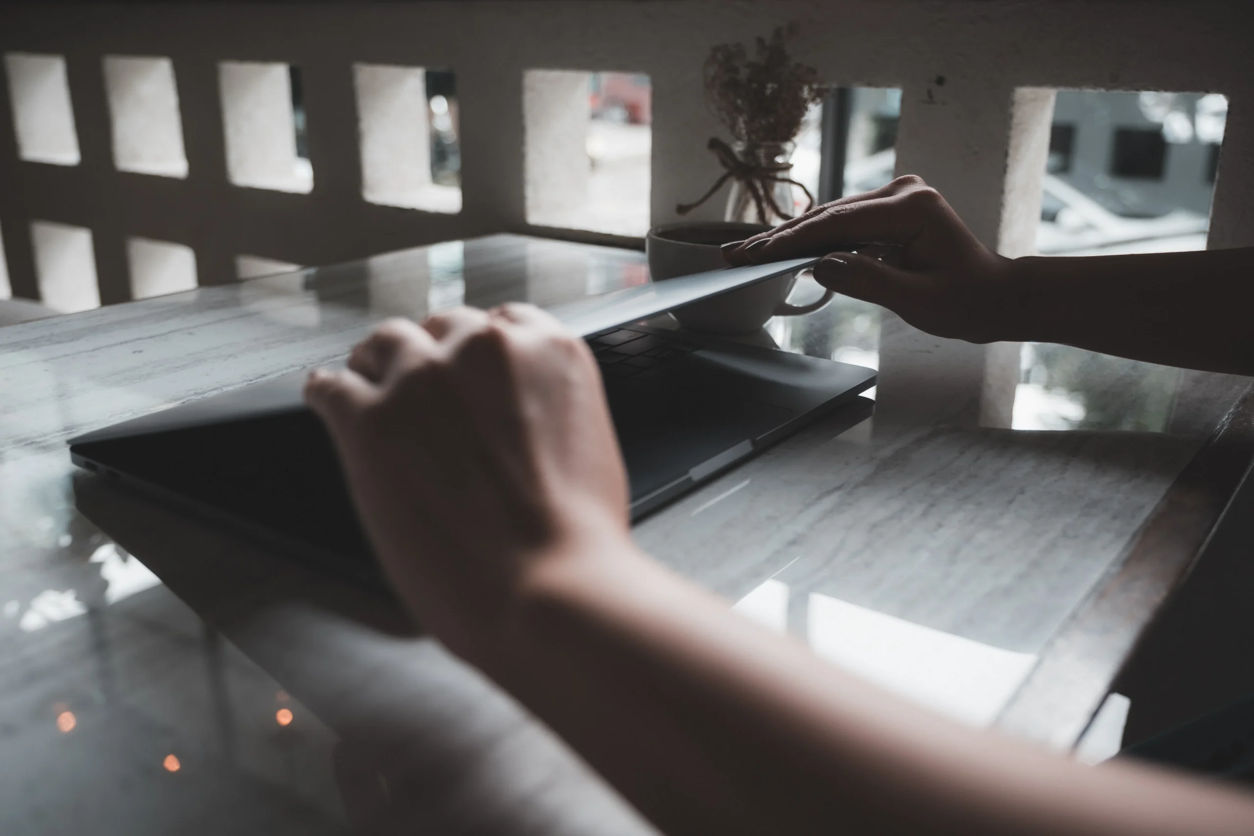 Close-up of hands closing a laptop on a marble table beside a coffee cup and dried flowers