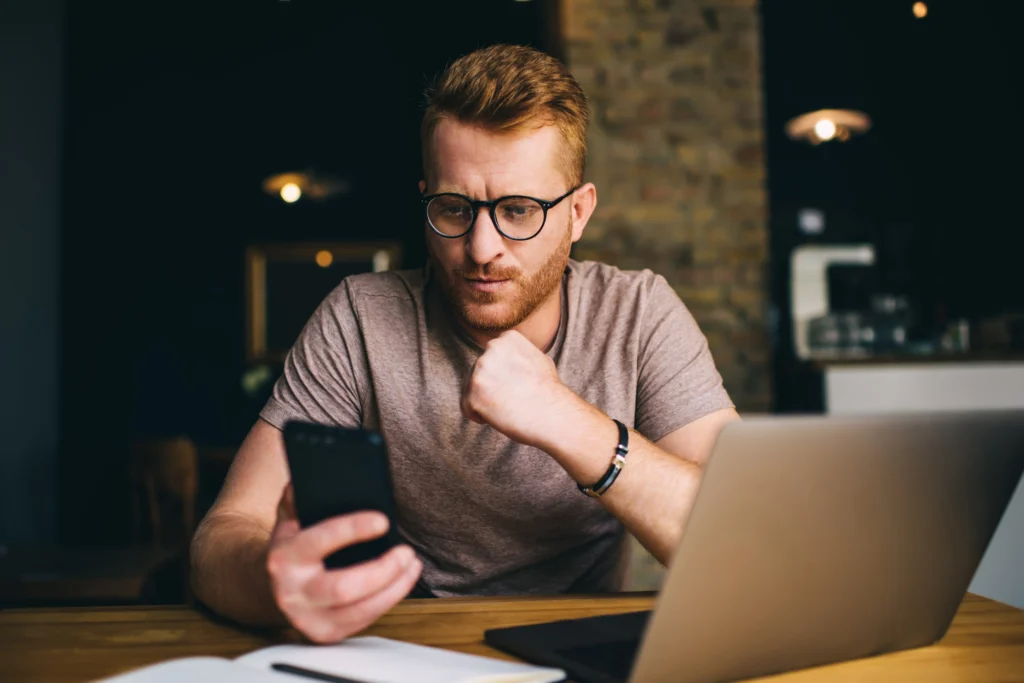 Man with glasses looking concerned while checking his phone beside an open laptop at night